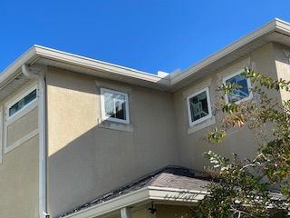 Beige stucco house with white-framed windows, gutters, and a clear blue sky.