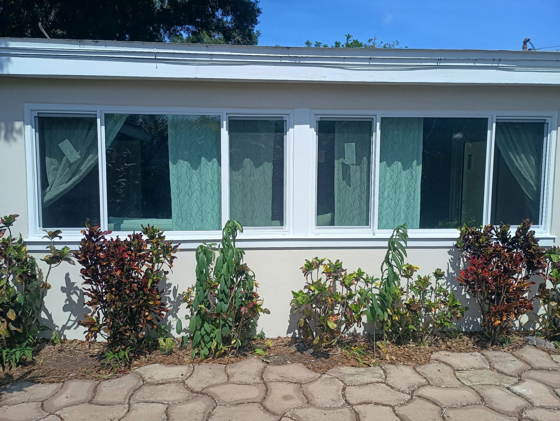 Four windowed building with white trim, green curtains, and small plants in front. Sunny day.