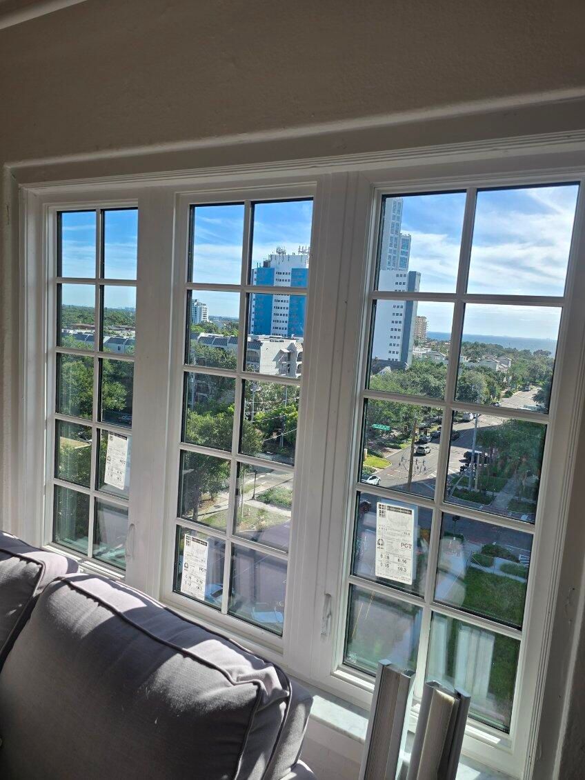 Three-paneled window with cityscape view. White frames, blue sky, buildings, trees, and partial grey sofa.