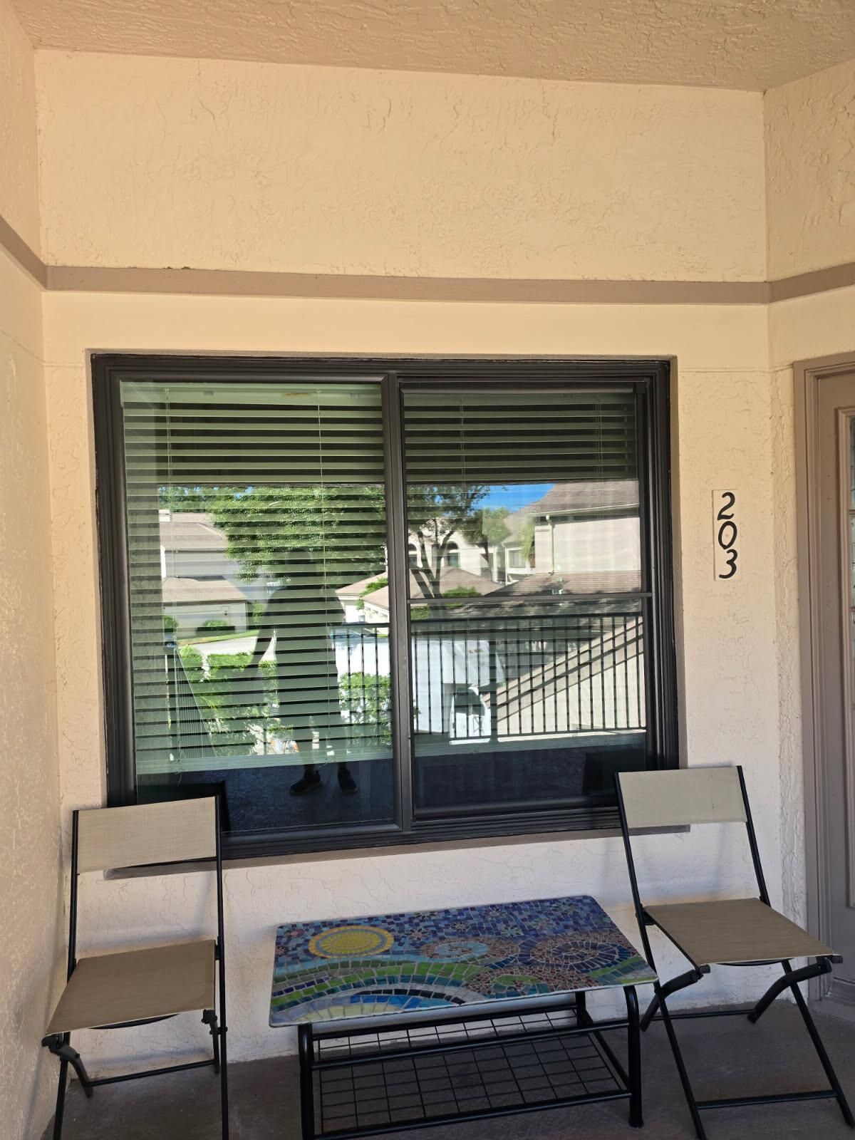 Balcony with chairs, a mosaic table, and a window with blinds overlooking a neighborhood.