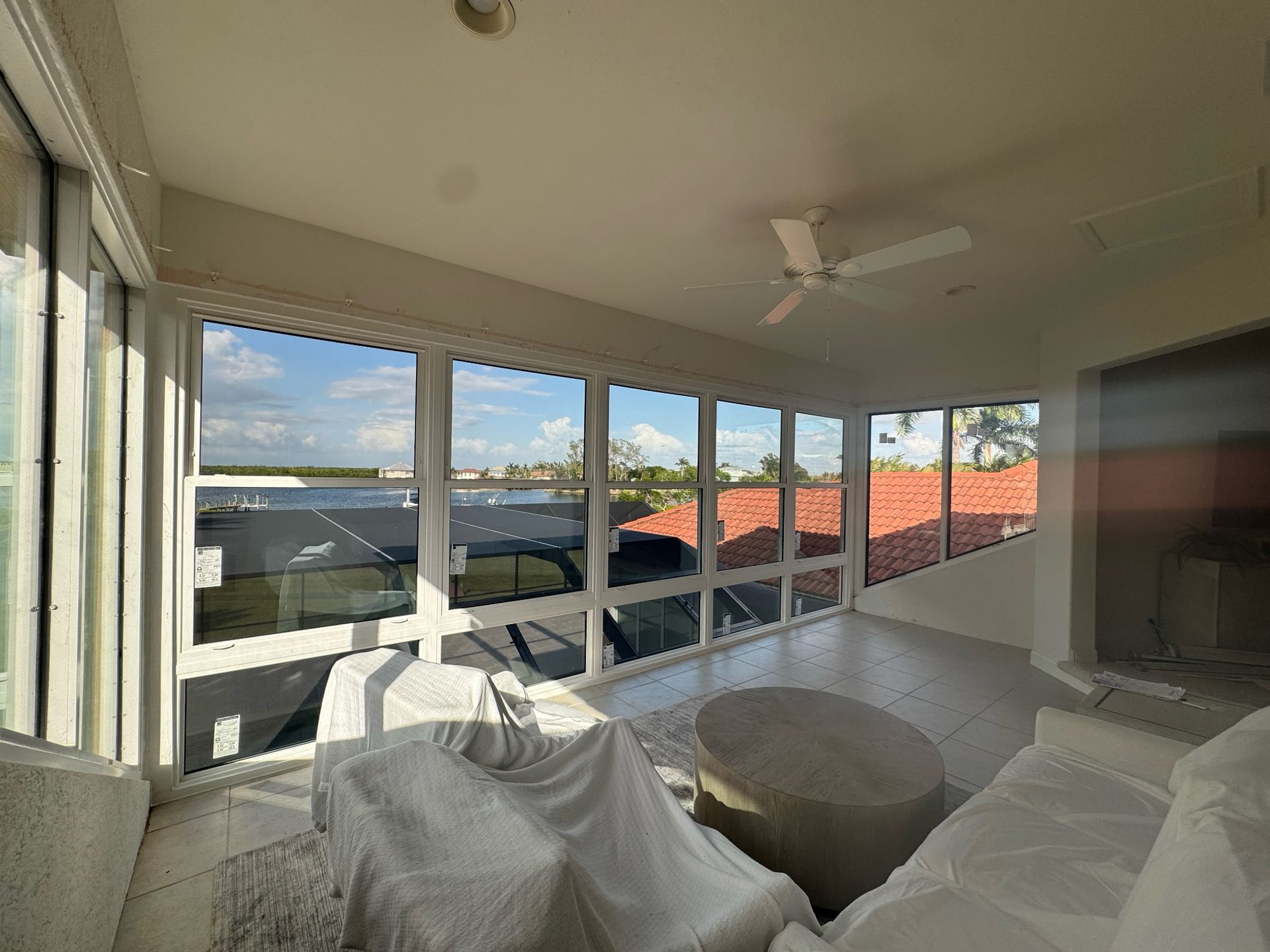 Sunroom with large windows overlooking a waterfront view, white furniture covered with protective sheeting.