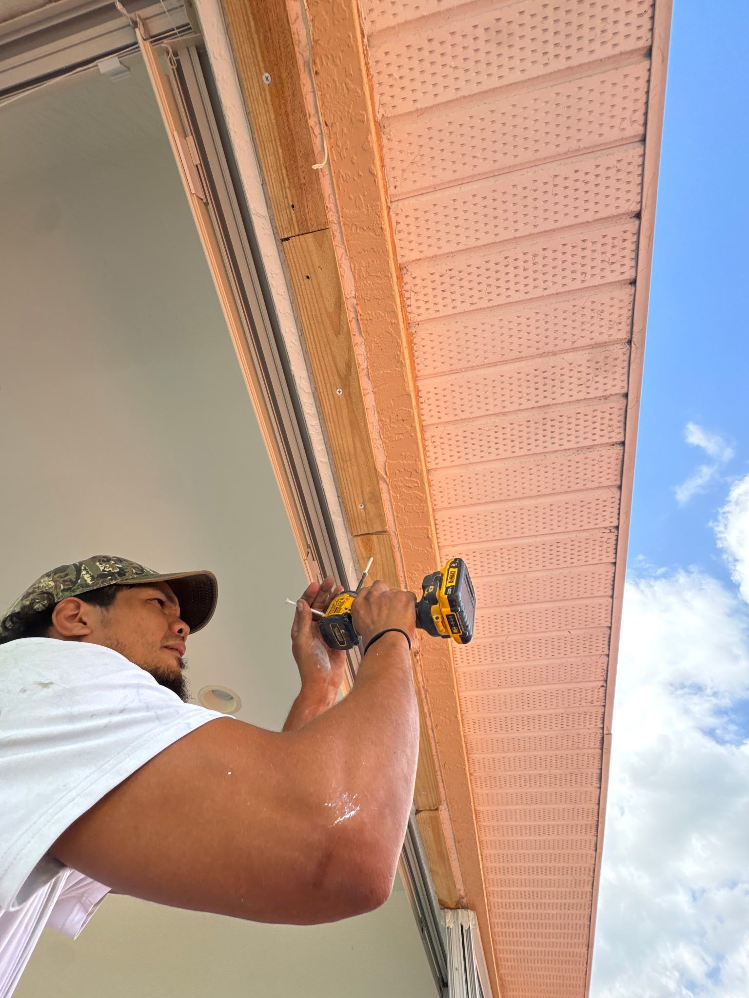 Person uses a power drill to install light pink siding under a beige overhang against a blue sky.