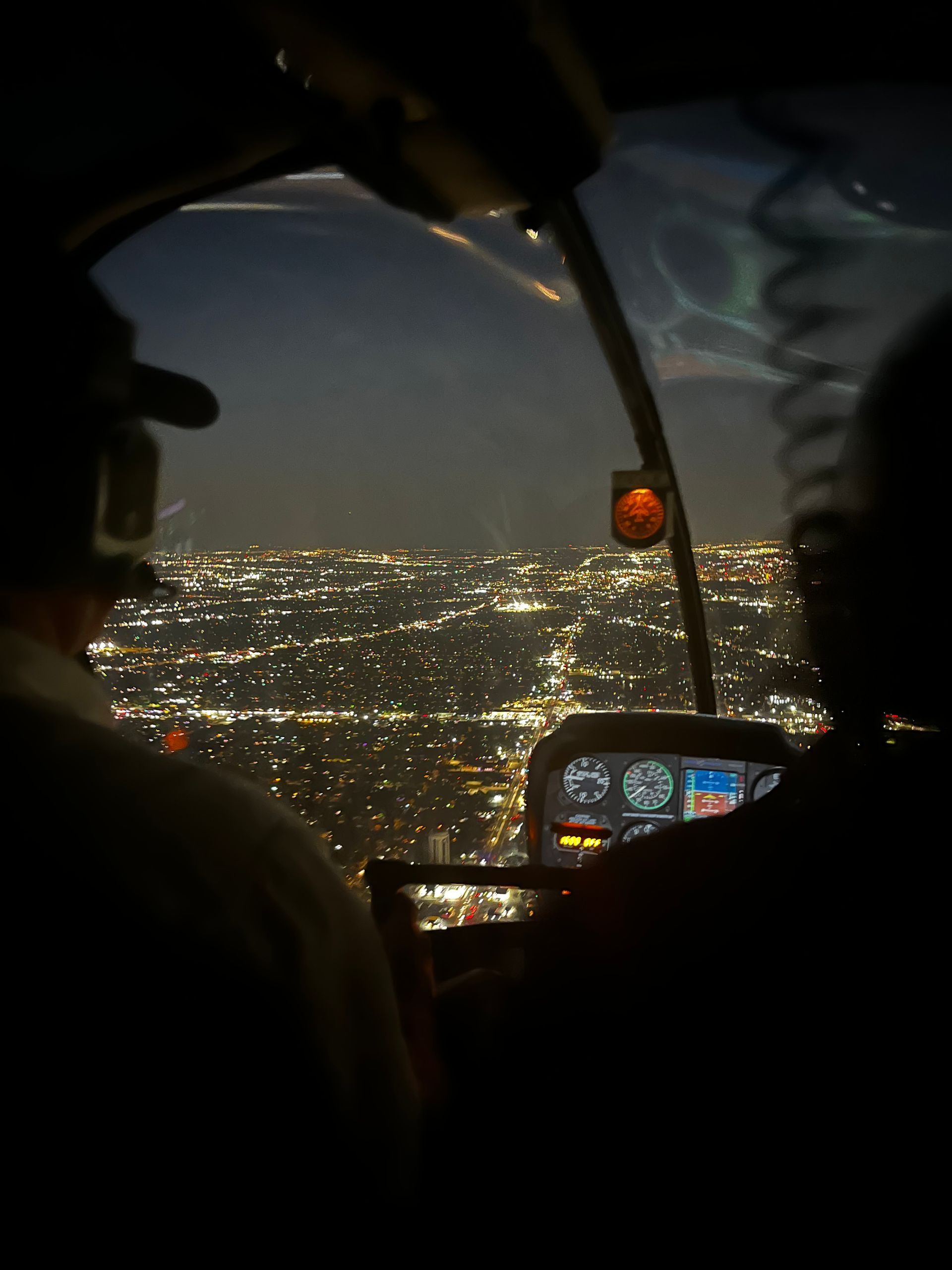 Night aerial view from a helicopter cockpit of a city's illuminated lights. Two people visible inside.