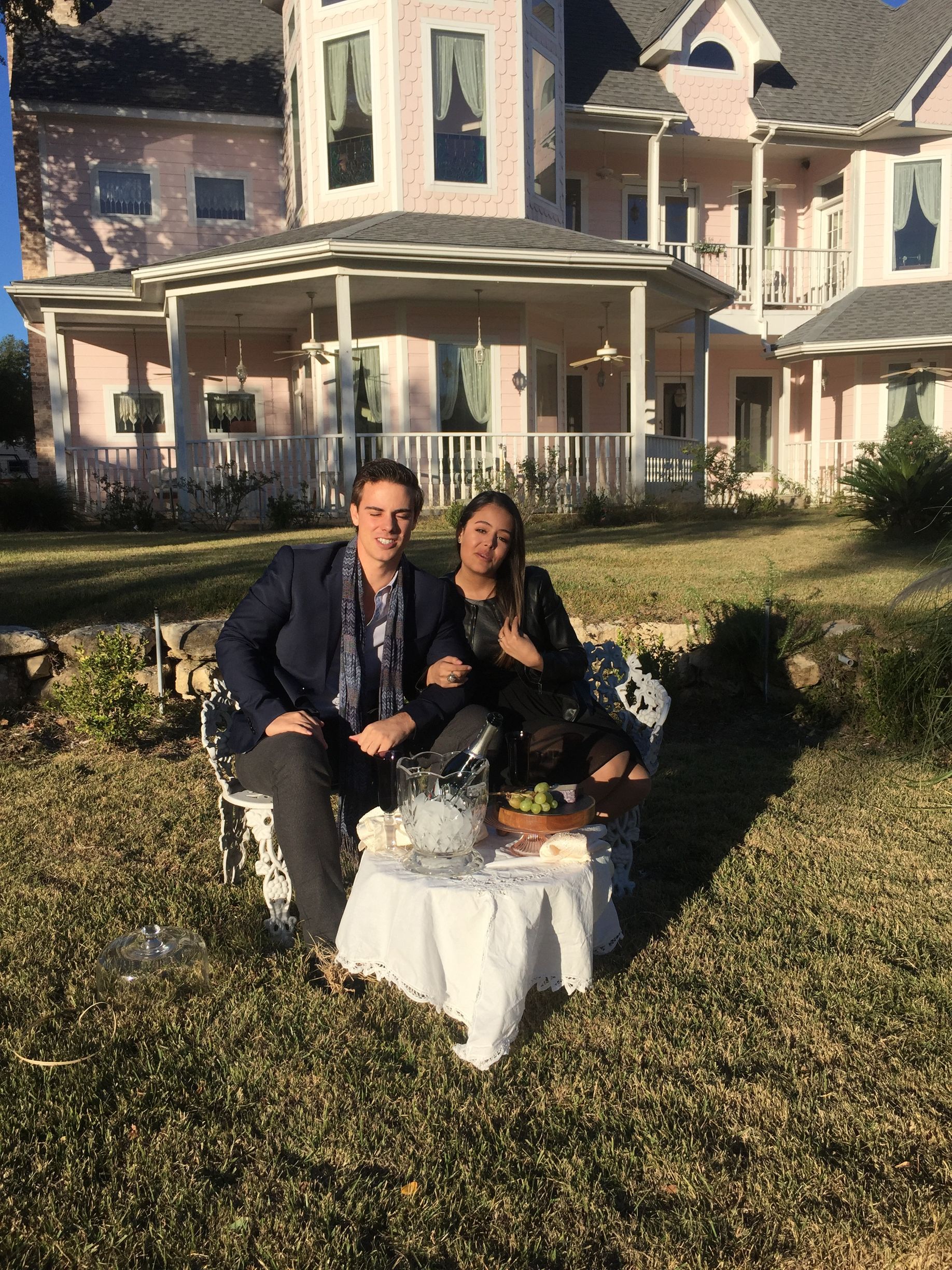 Couple sits outside a pink Victorian house. They are seated on a white table with refreshments.