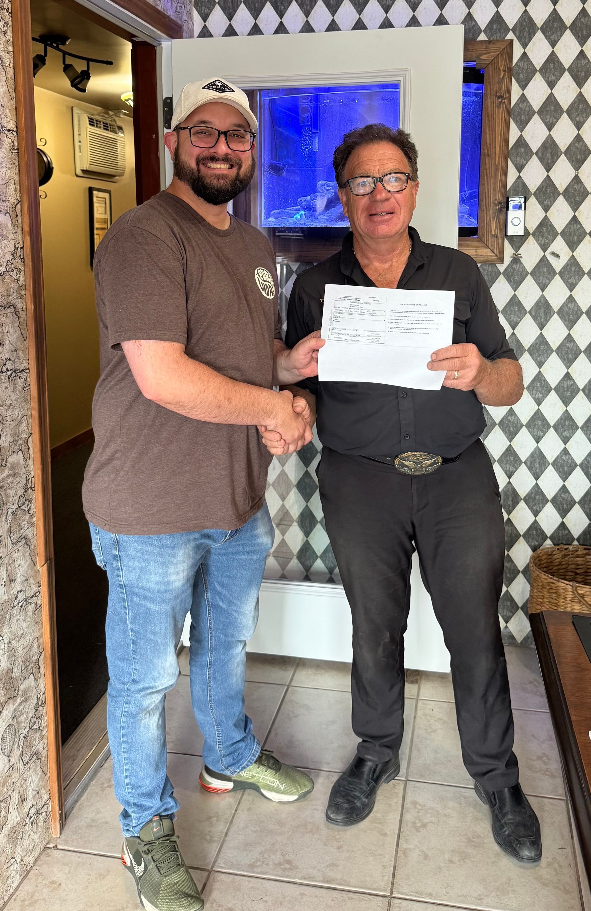 Two men shake hands, one holds a document. They stand inside, near a doorway and a decorative wall.