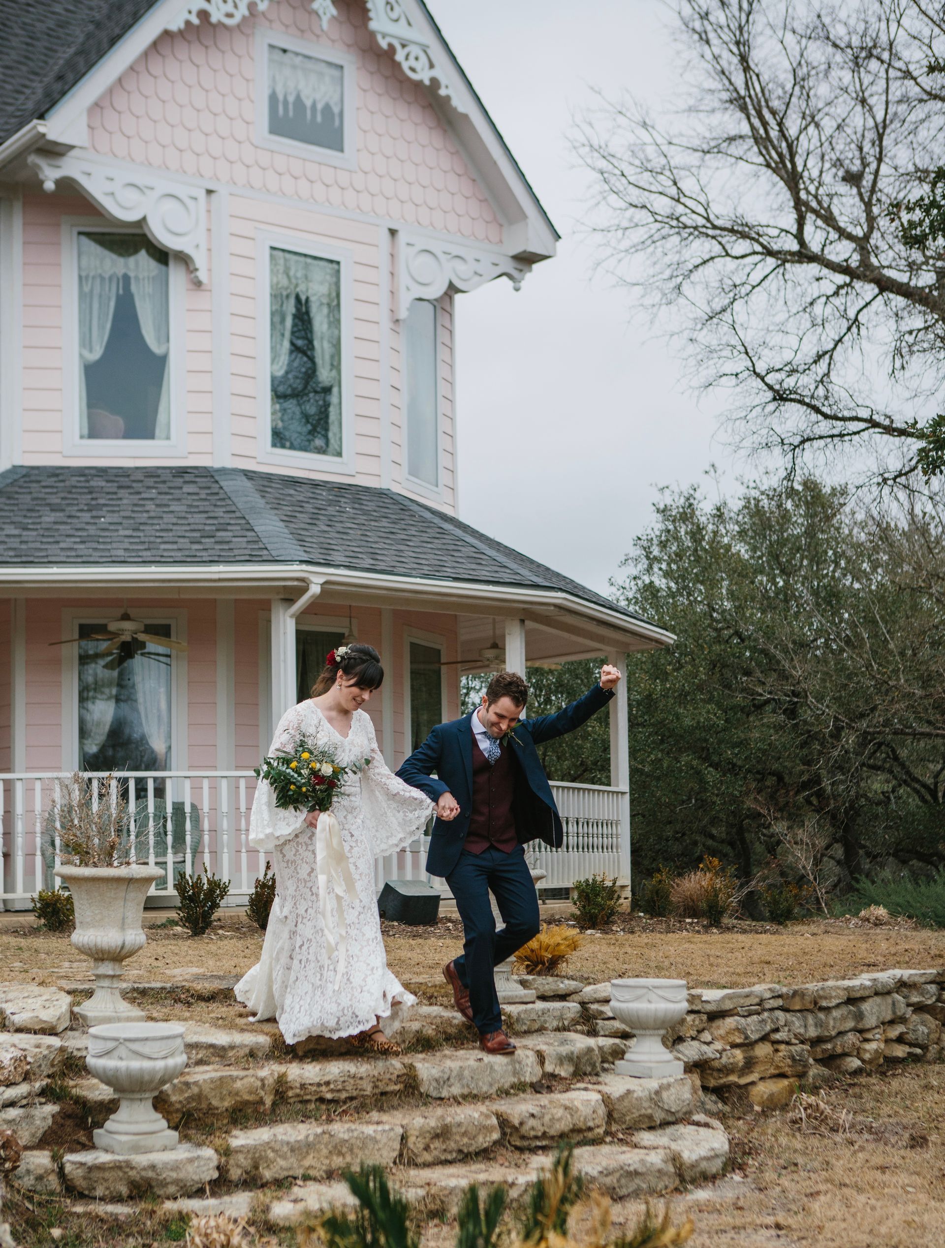Bride and groom exit a pink house after a wedding. Groom dances, bride holds bouquet. Cloudy day.