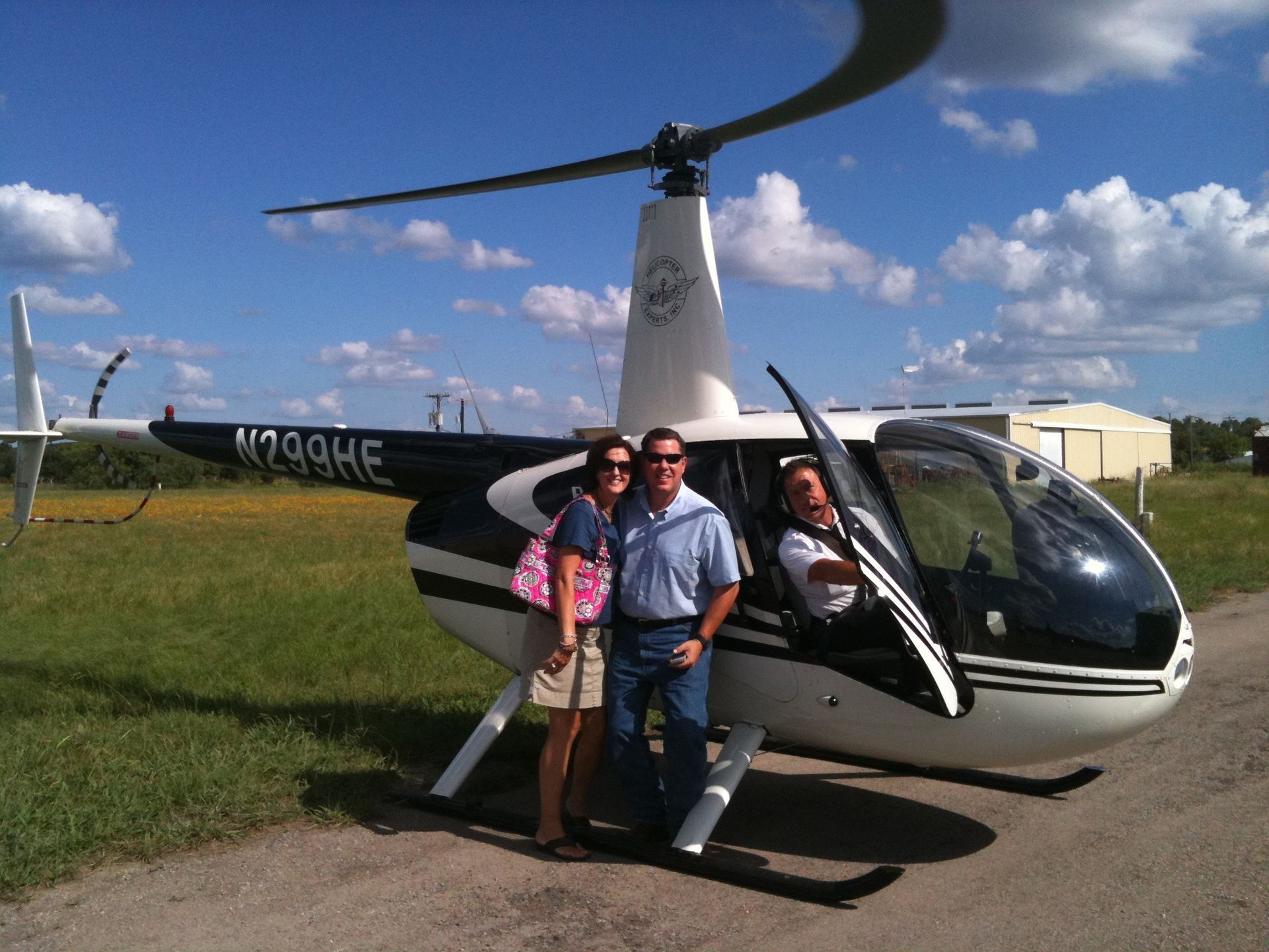 Couple poses with helicopter, pilot in cockpit, blue sky, and grass.