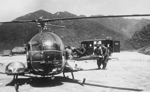 A medical helicopter on the ground, with medics and an ambulance in the background near mountains.