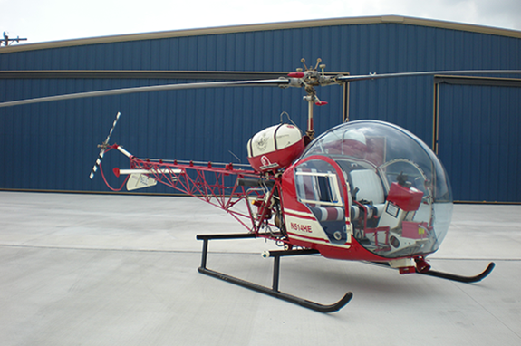 Red and white Bell 47 helicopter on a concrete surface, in front of a blue hangar.