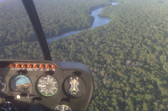View from a helicopter cockpit over a dense green forest and a winding river.