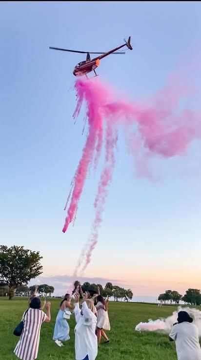 Helicopter releasing pink smoke for a gender reveal over a group of people on a grassy field.