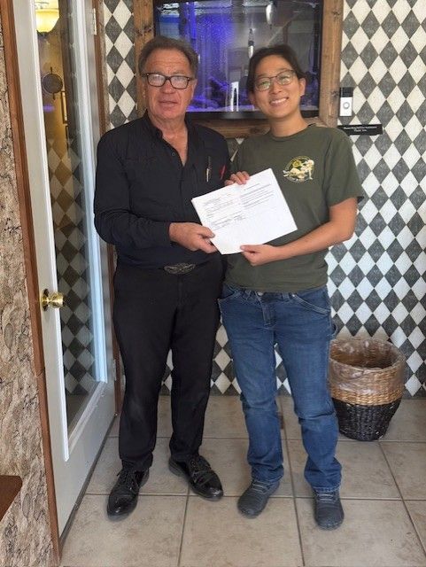 A person in black shirt hands a document to another person in green shirt and jeans, inside a building.
