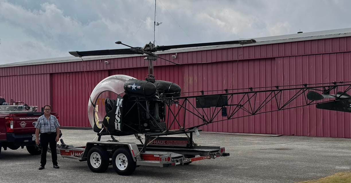 Man stands beside a black helicopter on a trailer in front of a red building.