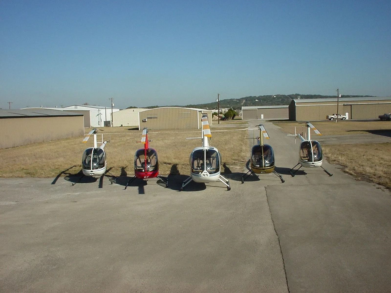 Five helicopters parked in a row on a tarmac under a clear, blue sky.
