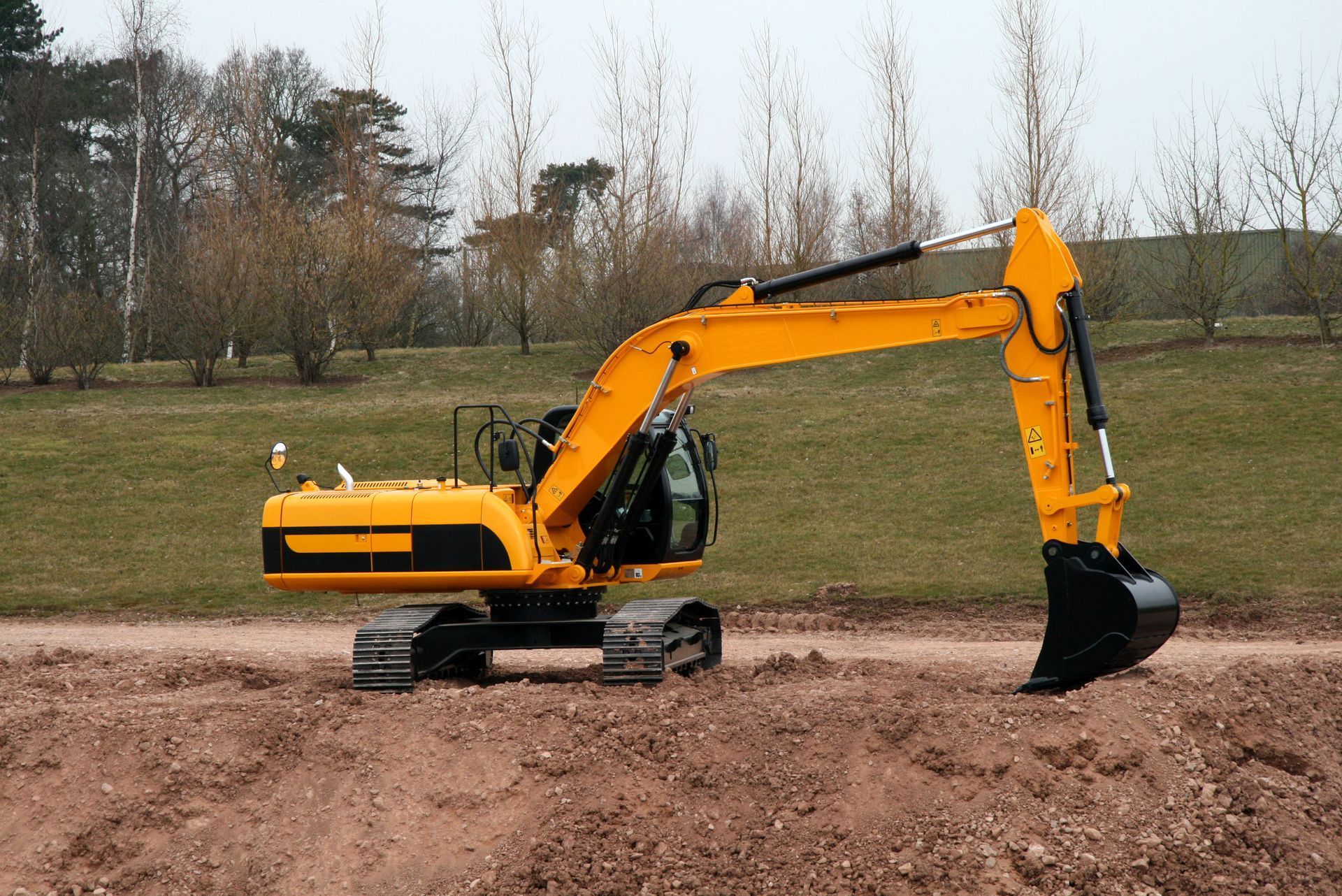 Yellow excavator on dirt path, near grassy hill and trees, arm extended with bucket.