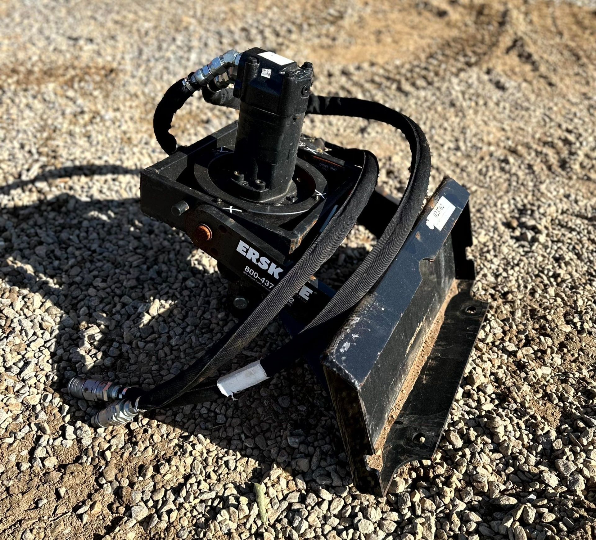 Black construction attachment on gravel, with hydraulic hoses.