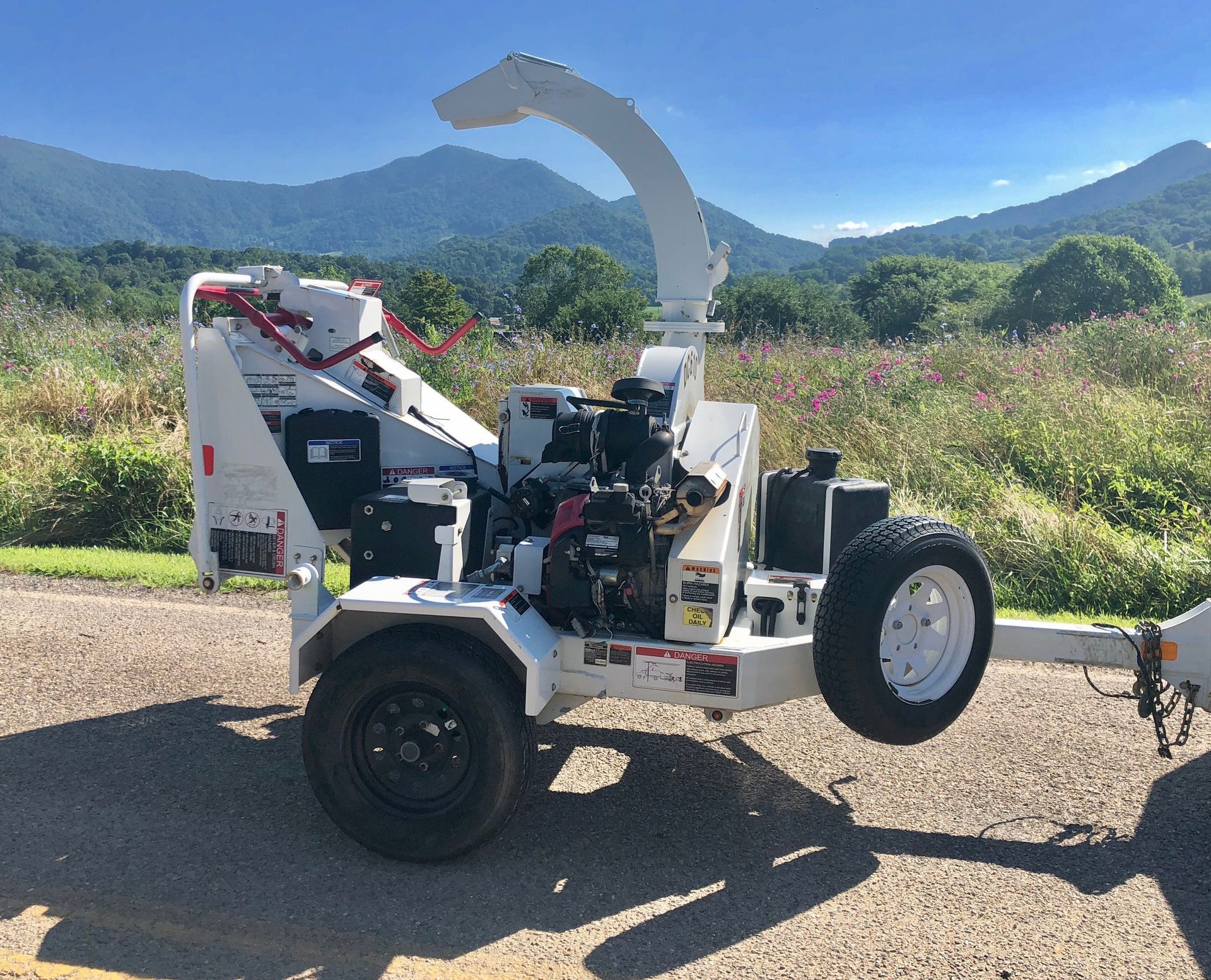 White wood chipper on a trailer, outdoors in front of mountains and trees, on a sunny day.