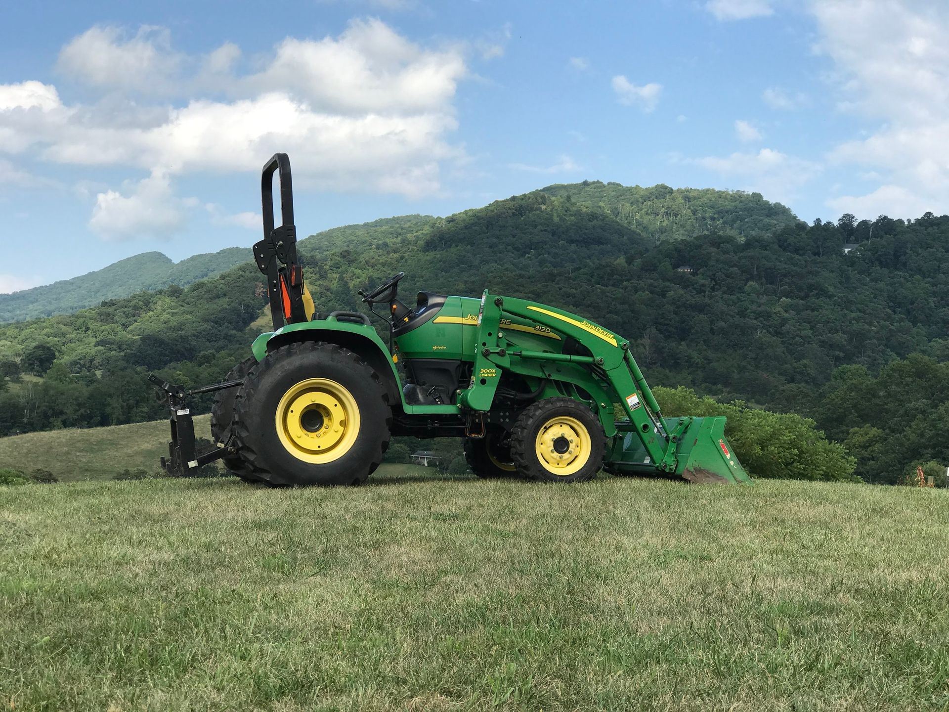 Green John Deere tractor on a grassy hill with mountains in the background under a blue sky.