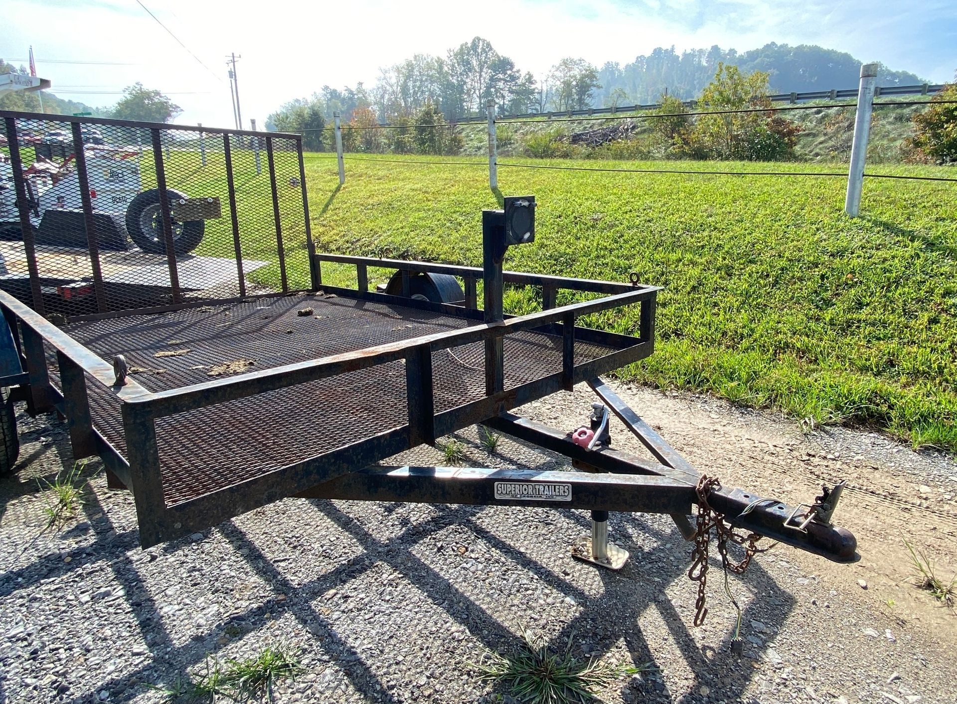 Black utility trailer on gravel, with wire mesh sides and back, parked outdoors.