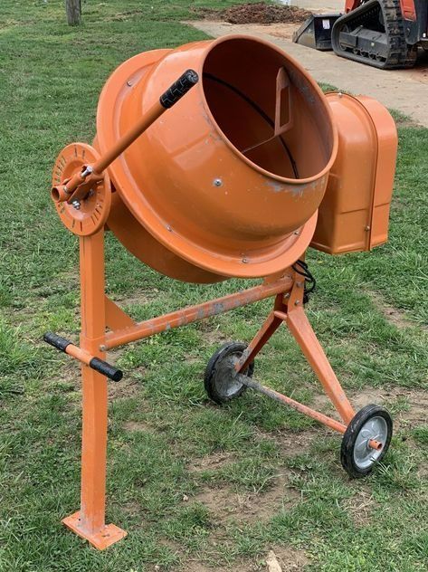 Orange cement mixer on a grassy surface. It has wheels and a handle for tilting.