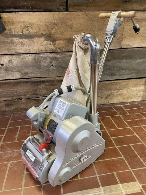 Floor sanding machine, gray metal with a handle, on a brick floor in front of wooden planks.
