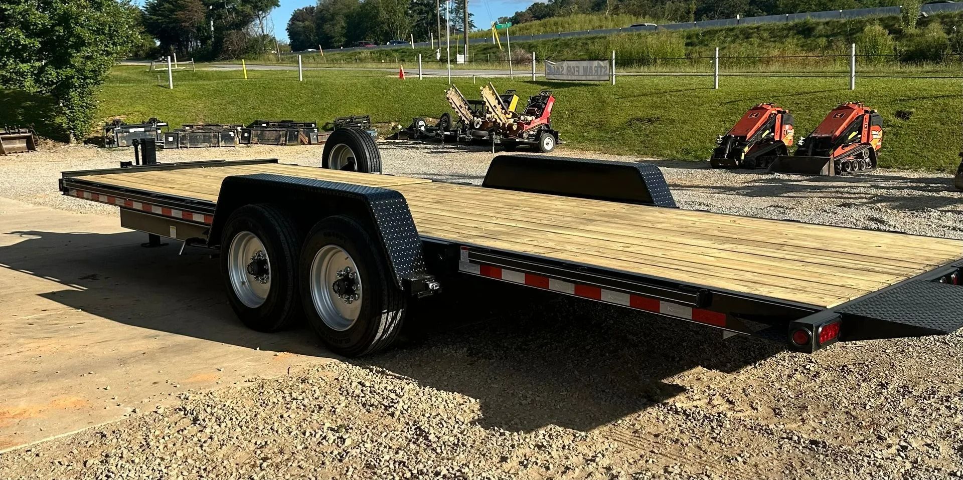 A flatbed trailer with two axles sits on gravel. Other vehicles are visible in the background.
