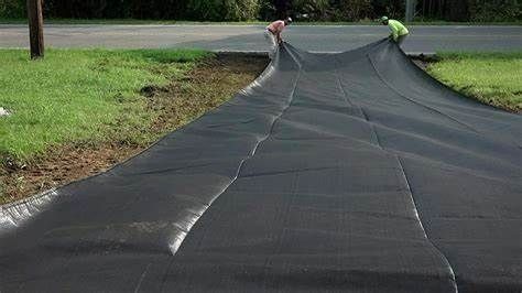 Two people installing black landscape fabric along a roadside curb, covering exposed dirt.