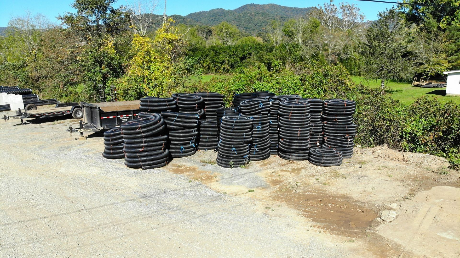 Stacks of black corrugated drain pipes on a gravel area, trees and a building in the background.