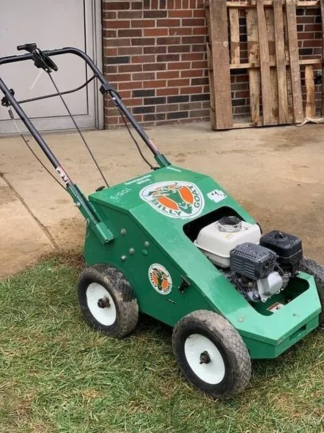 Green lawn aerator machine on grass. Brick building and pallets in background.