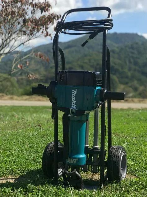 Makita demolition hammer on wheels, teal and black, set on grass, with a mountain backdrop.