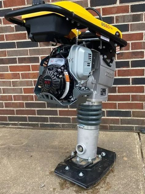 Yellow and black Wacker Neuson jumping jack compactor with engine and handle. On concrete near a brick wall.