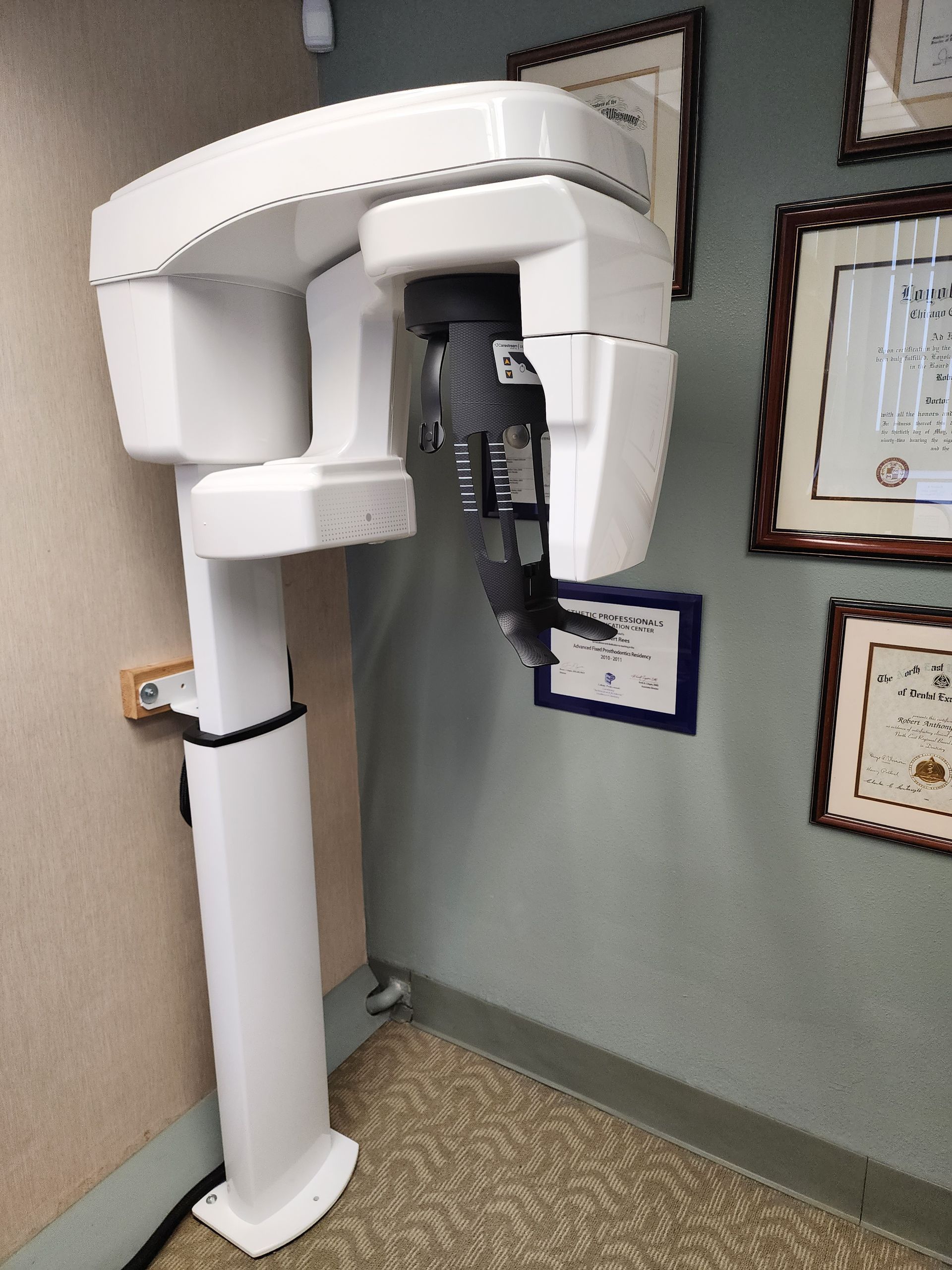 White dental X-ray machine in a clinic corner beside framed certificates.