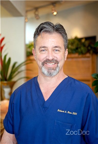Smiling man in blue medical scrubs standing indoors with a blurred clinic background