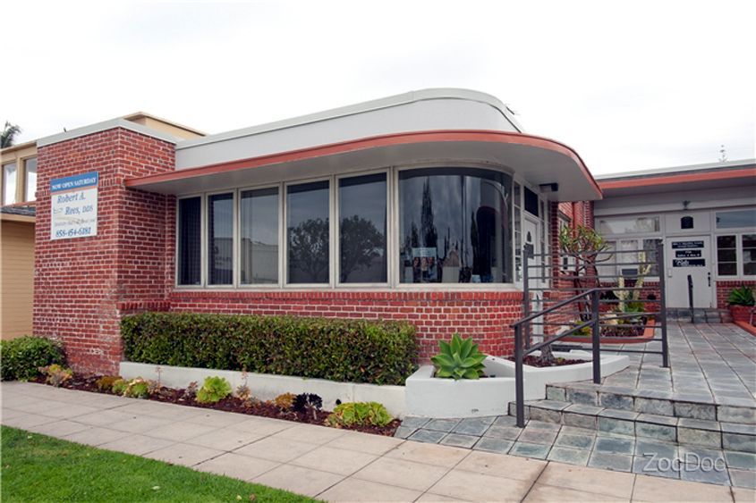 Brick building entrance with large windows, landscaped bushes, and steps leading to a doorway