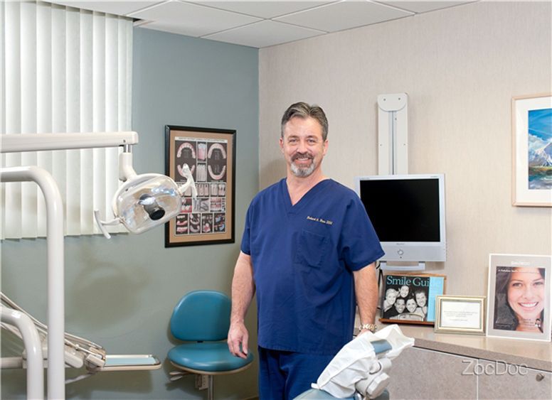 Dentist standing in a bright exam room with dental chair, monitor, and treatment equipment