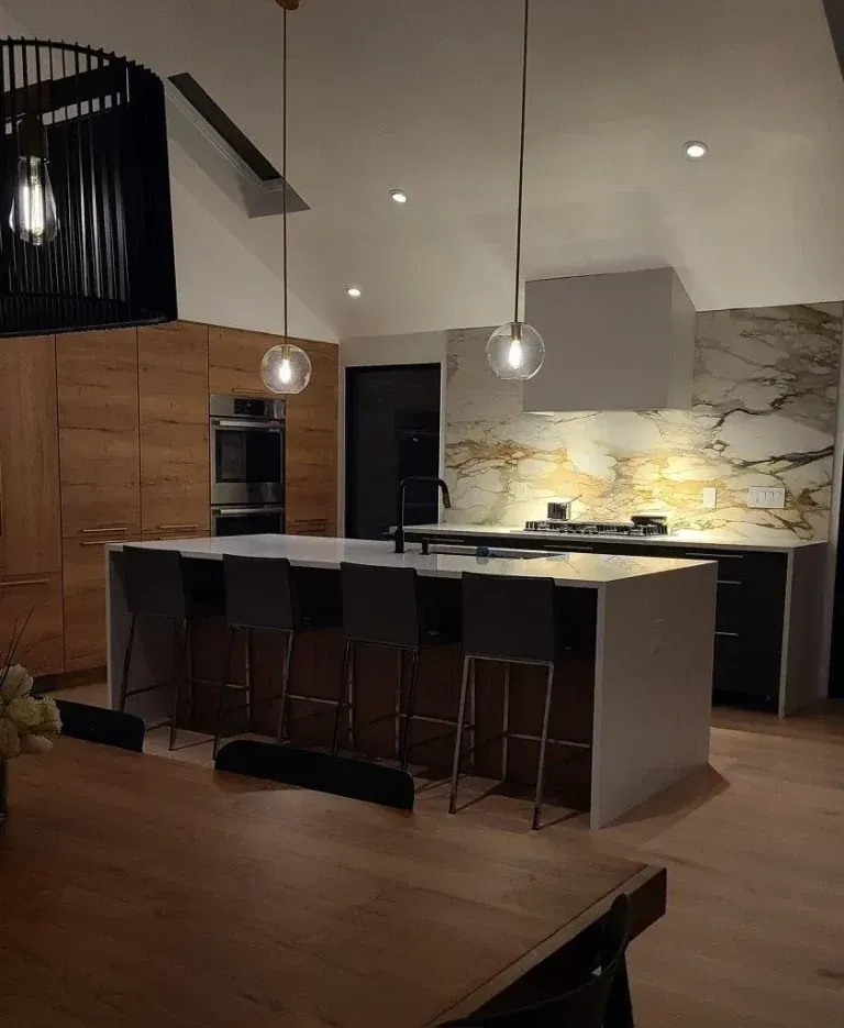Modern kitchen with large island, pendant lights, bar stools, and a marble backsplash at night