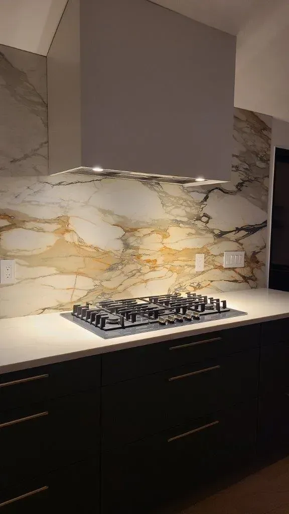 Modern kitchen with marble backsplash, dark cabinets, and a gas cooktop under a stainless hood.