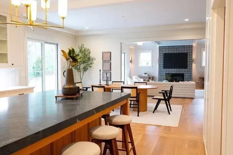 Bright modern kitchen opening to a living room with island stools, wood floors, and a large TV fireplace.