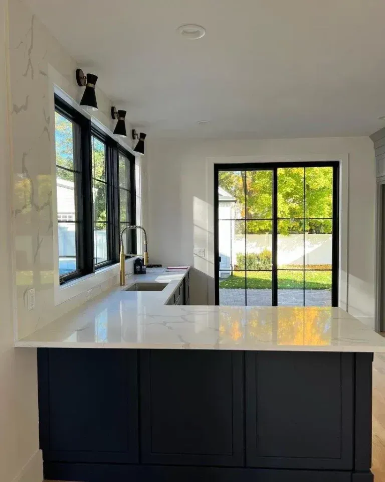 Bright modern kitchen with white island, black cabinets, and large windows overlooking greenery.