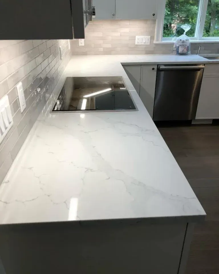 Modern kitchen with white marble countertop, built-in cooktop, stainless dishwasher, and window view.