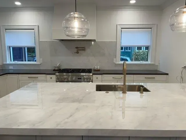 Modern kitchen with white marble island, stainless range, pendant lights, and two windows.