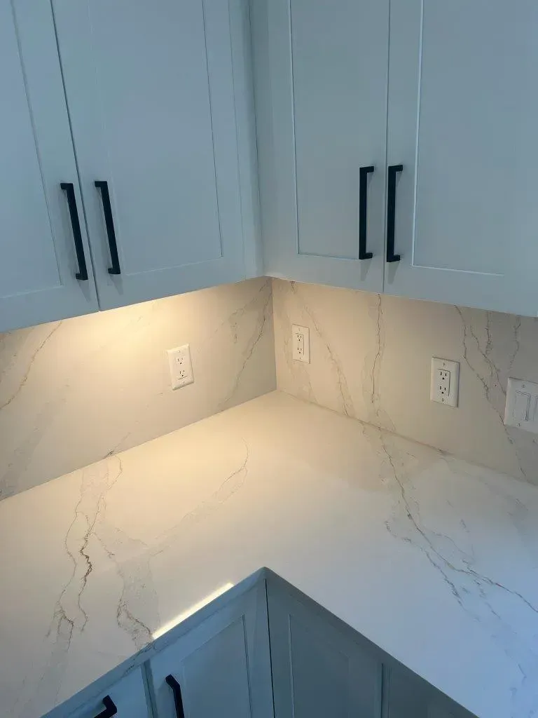 White kitchen corner with marble-look countertop, light gray cabinets, black handles, and under-cabinet lighting