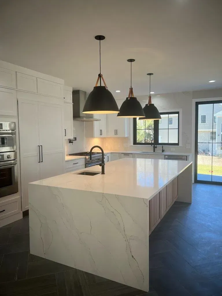 Modern kitchen with large marble island, black pendant lights, white cabinets, and window view to outdoors