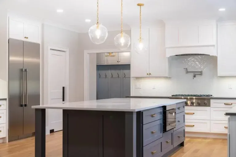 Bright white modern kitchen with a dark island, stainless fridge, and hanging pendant lights.