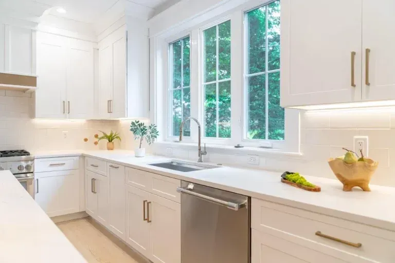 Bright white modern kitchen with gold handles, large window, sink, and a dishwasher on the counter wall