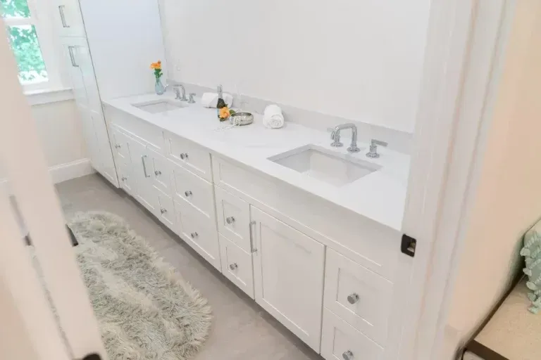 Bright white bathroom vanity with two sinks, drawers, and a shag rug beside a window.