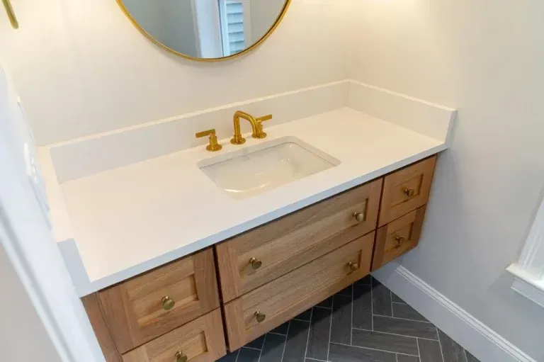 White bathroom vanity with wood drawers, gold faucet, round mirror, and dark herringbone tile floor