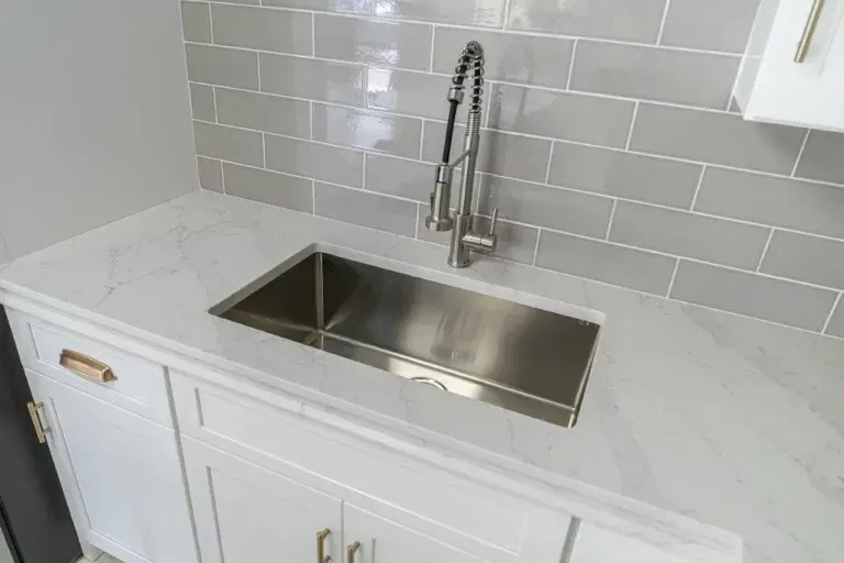 White kitchen counter with stainless steel farmhouse sink and chrome faucet against gray tile backsplash