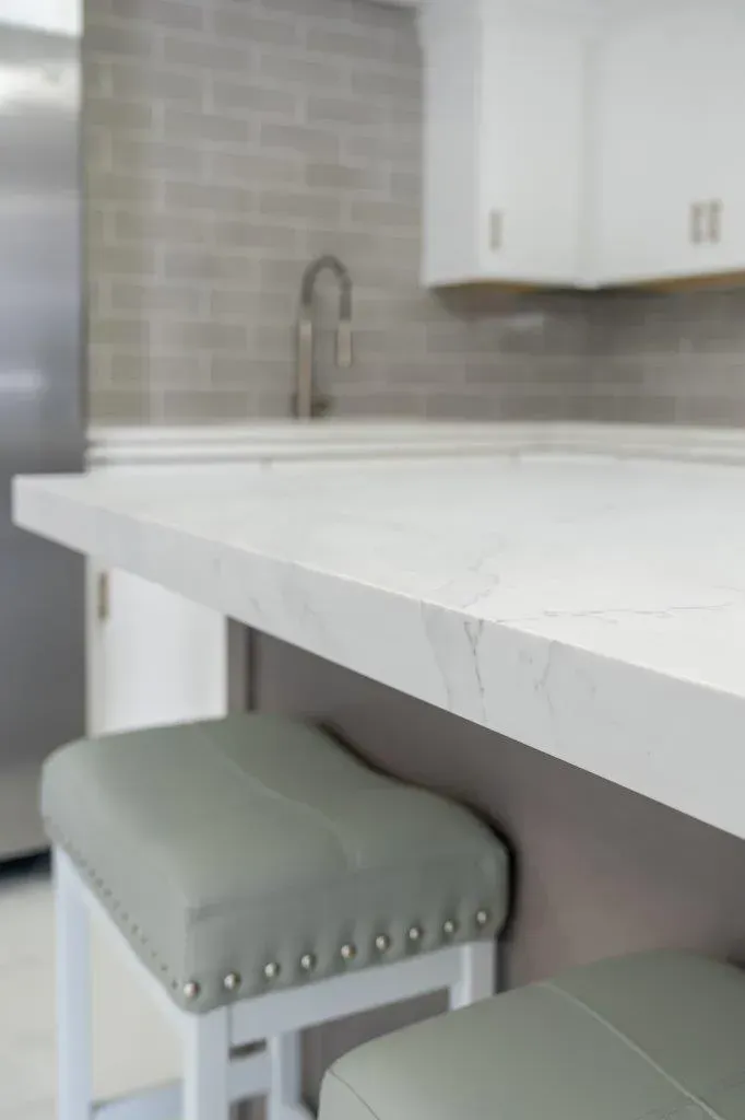 Kitchen island with white countertop, pale gray tile backsplash, cabinets, and two mint-green stools