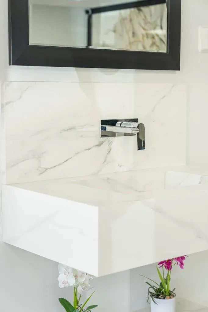Modern white bathroom sink with chrome faucet, marble-look wall, and black-framed mirror above.